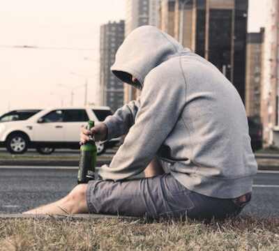 man with a hoodie on his head on a bright day, sitting alone with a beverage in his hand on the end of a road