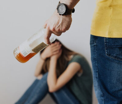 man with an alcohol bottle going towards a woman sitting on the floor, covering herself in self-defense. The rage and aggressiveness from alcohol.