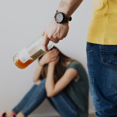 man with an alcohol bottle going towards a woman sitting on the floor, covering herself in self-defense. The rage and aggressiveness from alcohol.