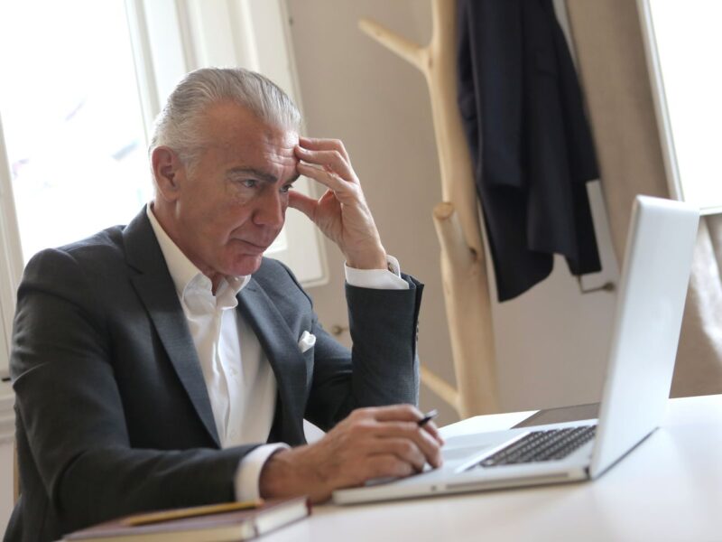 Older man holding his head and working on a laptop