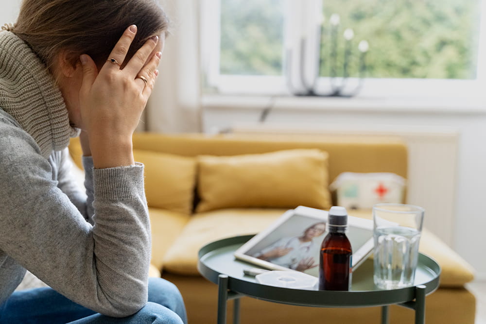 Middle‑aged person in grey sweater having online telehealth consultation at home, medicine bottle and glass of water on coffee table in foreground, blurred female doctor on tablet screen.