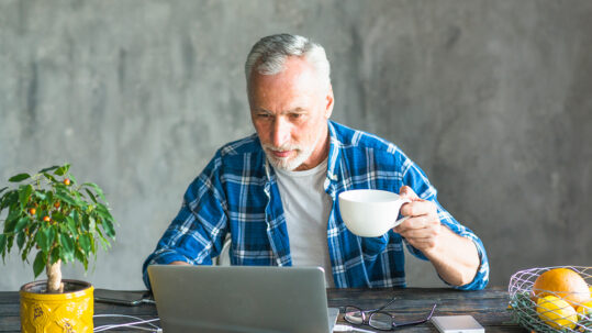 Senior man working remotely at home, older man in blue plaid shirt using laptop and drinking coffee at wooden desk, relaxed home office routine with plant and fruit on table.