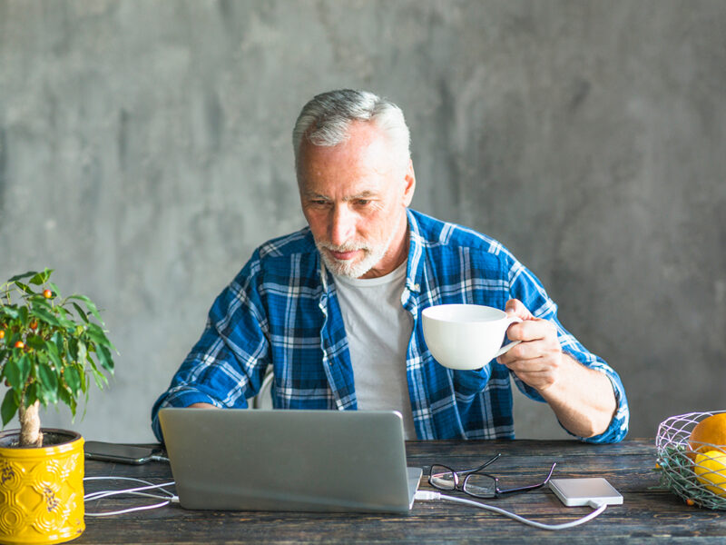 Senior man working remotely at home, older man in blue plaid shirt using laptop and drinking coffee at wooden desk, relaxed home office routine with plant and fruit on table.