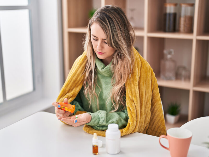Woman wrapped in yellow blanket taking medication at home, sick woman holding pill bottle and tablets at white table, cold and flu treatment concept.