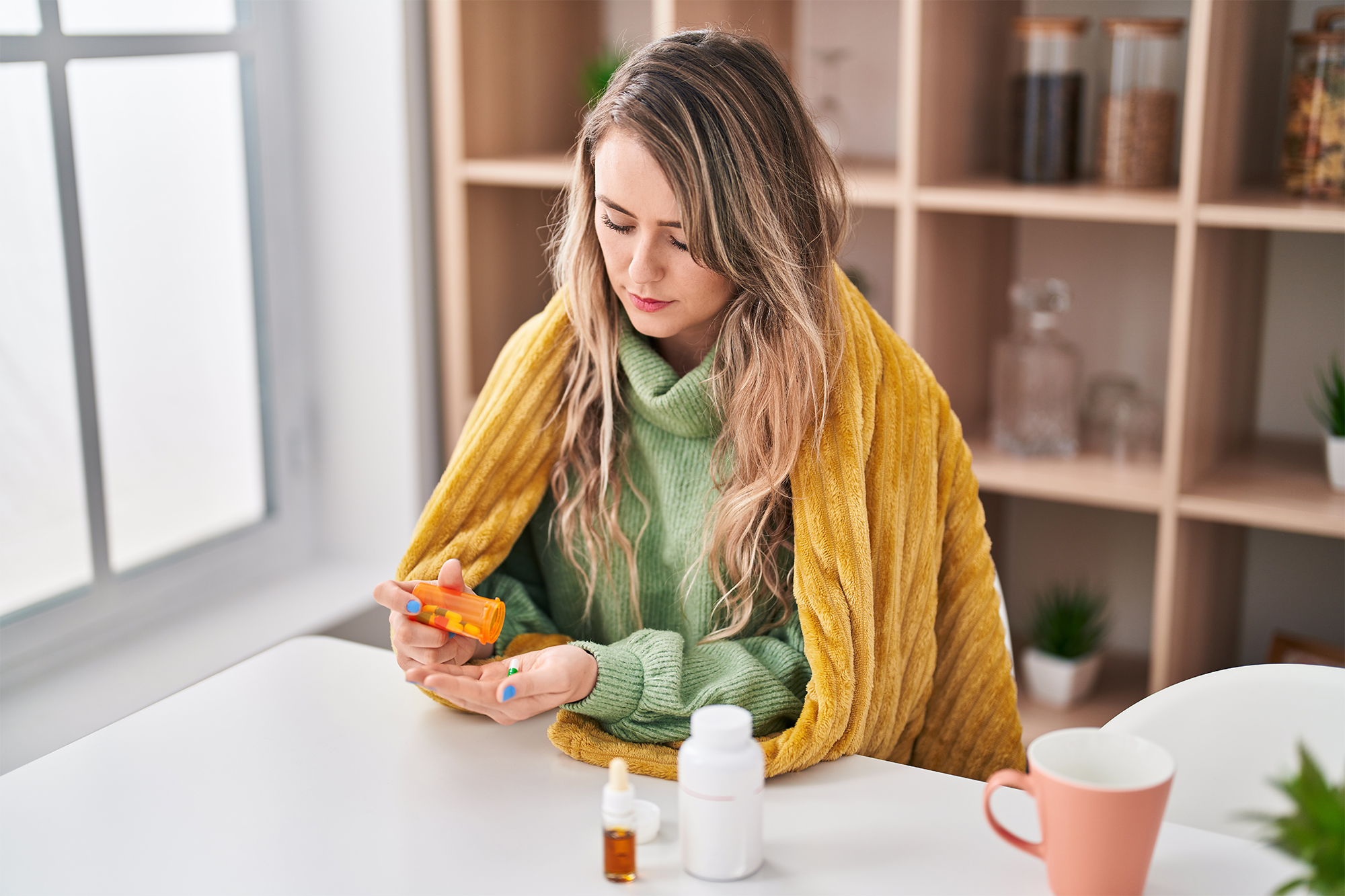 Woman wrapped in yellow blanket taking medication at home, sick woman holding pill bottle and tablets at white table, cold and flu treatment concept.