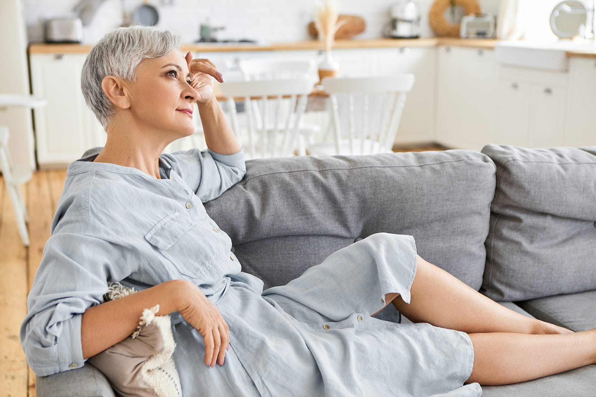 Side view of attractive stylish european female pensioner in elegant dress sitting barefooted on comfortable couch, having thoughtful look, daydreaming. People, retirement, mature age and lifestyle